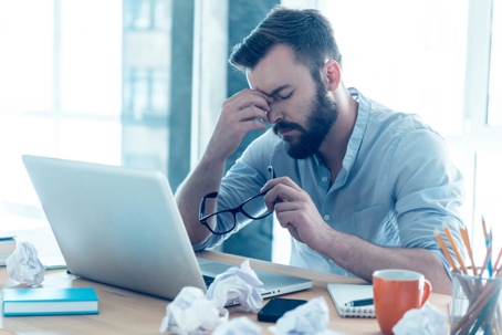 Frustrated young beard man massaging his nose and keeping eyes closed while sitting at his working place in office.